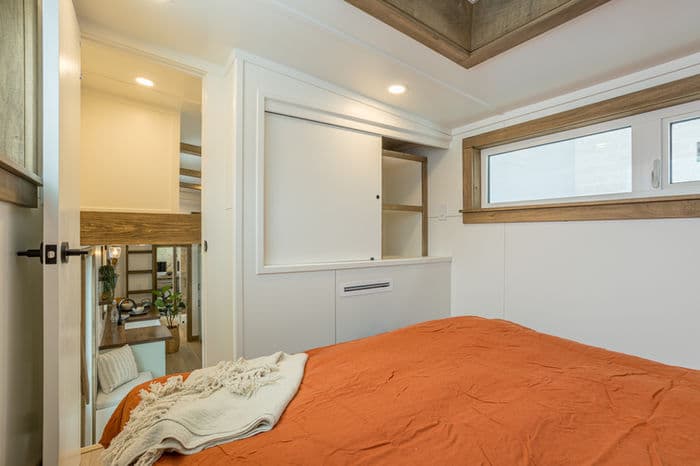 A cozy tiny house bedroom with an orange bedspread and beige throw, featuring built-in storage with sliding doors, a window above the bed, and a view into a brightly lit, modern living area.