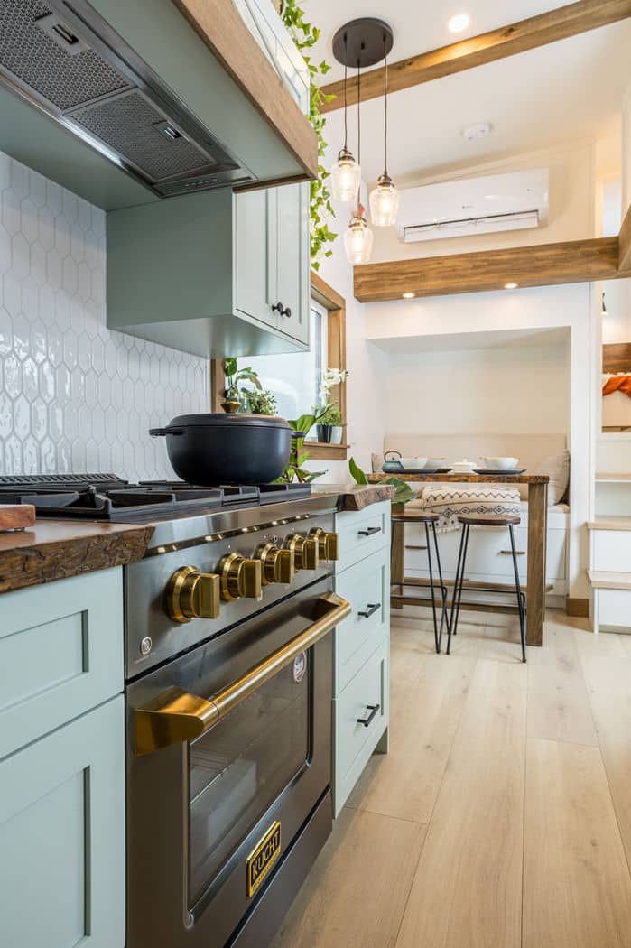 Modern tiny house kitchen with mint green cabinets, gold-accented oven, white hexagon tile backsplash, wood countertops, and open shelving with plants; a cozy dining area with a table and bench sits in the background.