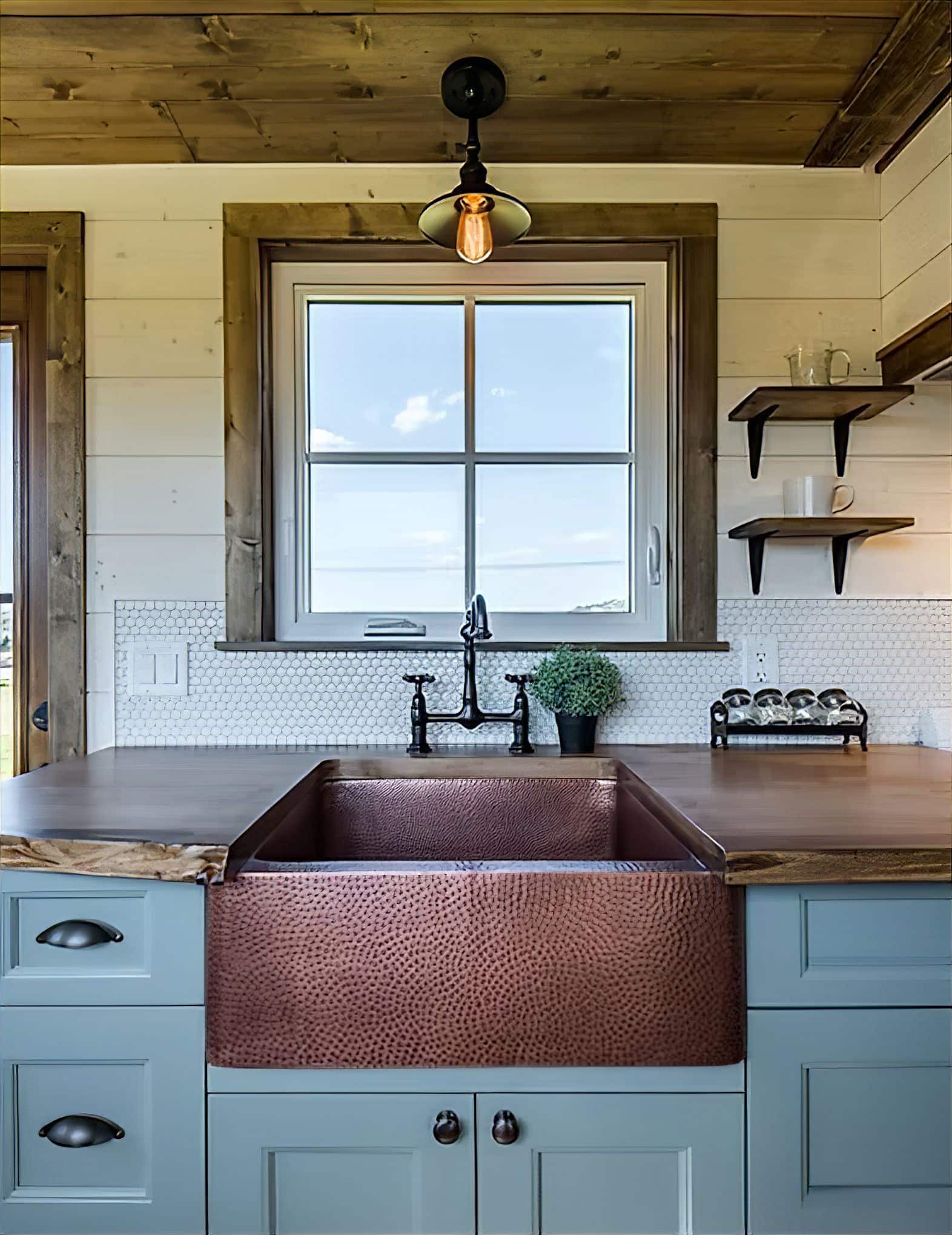 A farmhouse kitchen sink with a hammered copper finish is set in blue cabinetry, beneath a window. Wooden countertops, white tile backsplash, and open shelves complete the rustic decor, making it perfect for a tiny house kitchen.