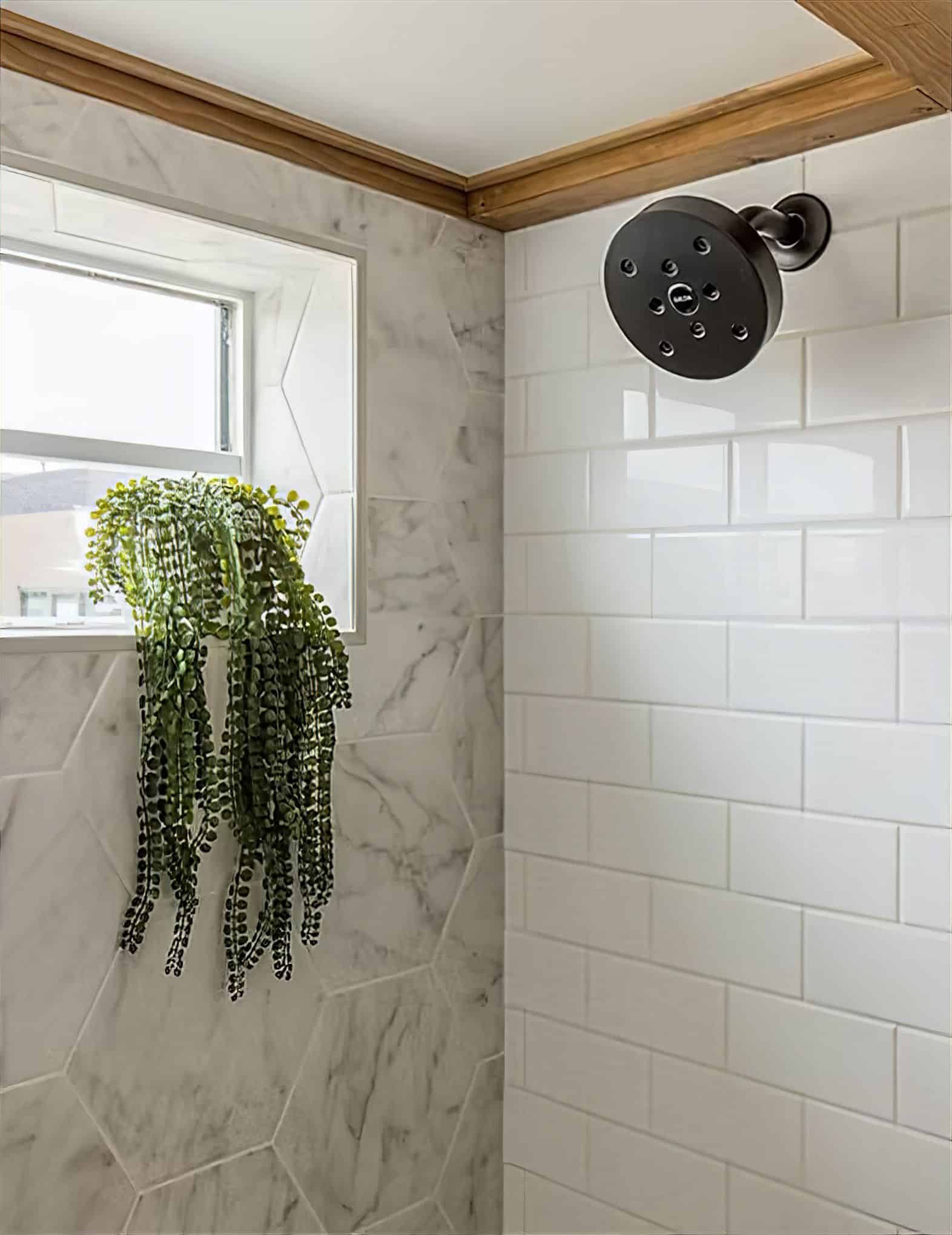 A tiny house shower with white subway tile walls, a round showerhead, and a small window holding a trailing green plant. The window is framed with marble-look hexagon tiles and wood trim.