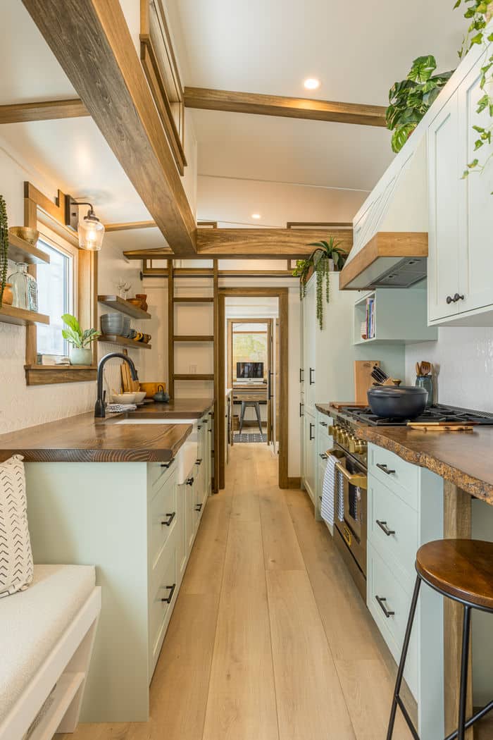 A narrow, modern tiny house kitchen with light green cabinets, wooden countertops, open shelves with plants, a black stove, and natural light from a window. Light wood floors and a cozy, efficient design maximize every inch of space.