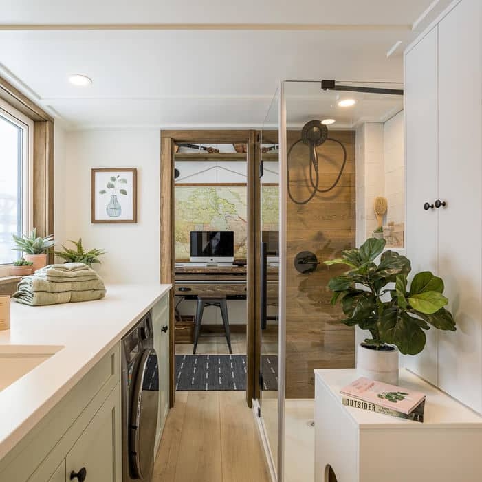 Modern tiny house bathroom with a glass shower, indoor plants, stacked towels, a washing machine, and a workspace with a computer and map visible through a doorway. Natural light streams in from a large window.