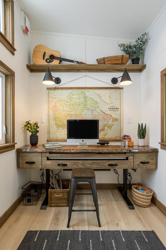 A cozy tiny house home office with a rustic wooden desk, computer, metal chair, plants, and a large map of Canada on the wall. Above the map, a shelf holds a guitar, basket, and decor as natural light pours in through the windows.