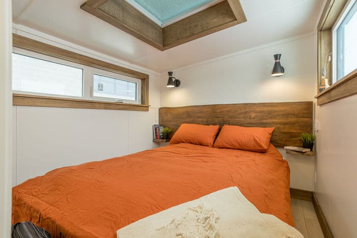 A cozy tiny house bedroom with an orange bedspread, wooden headboard, small built-in shelves, wall-mounted reading lights, windows on two sides, and a skylight in the ceiling.
