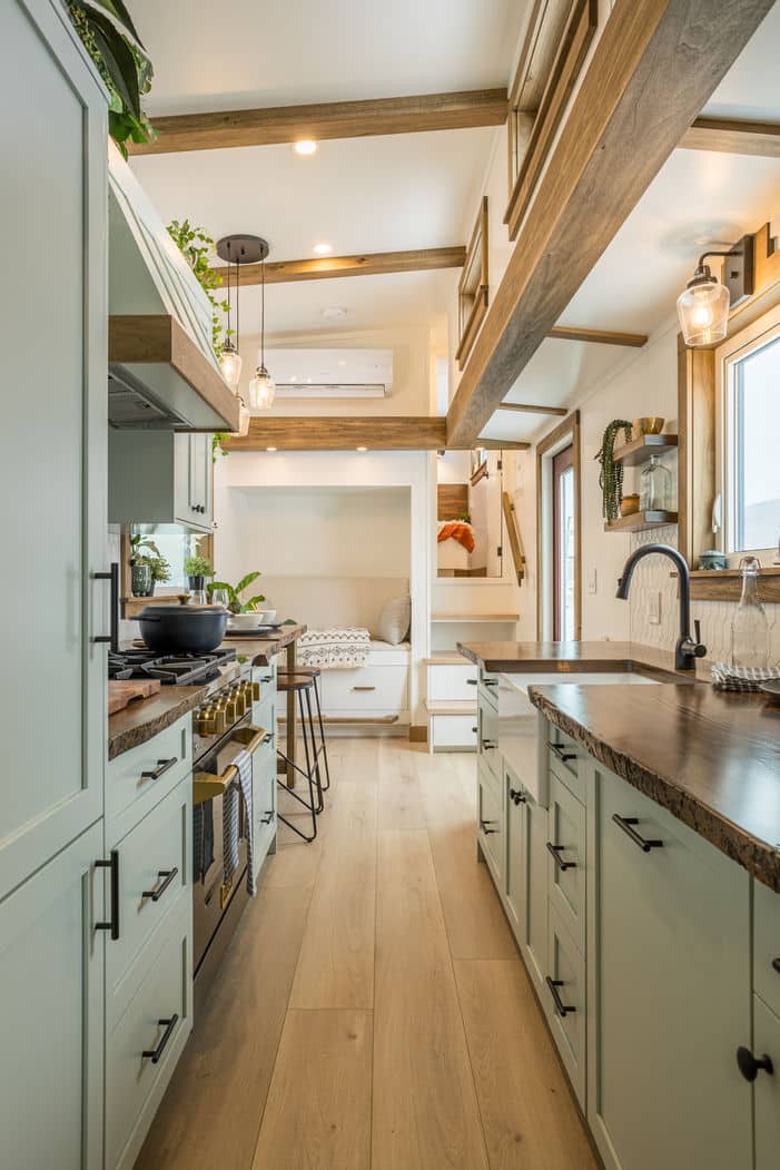 Bright, modern tiny house kitchen with light wood floors, sage green cabinets, stone countertops, and stainless steel appliances. A cozy loft and sitting area complete this inviting tiny house space, filled with ample natural light.