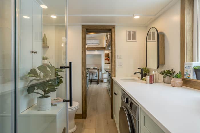 A modern, narrow bathroom in a tiny house features light wood floors, a washer and dryer, white cabinets, potted plants, a round mirror, and a view into the dining area beyond a wooden doorway.