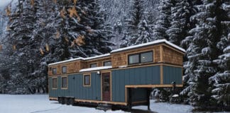 A blue tiny house with wooden trim sits on wheels in a snowy clearing, surrounded by tall, snow-covered evergreen trees and mountains in the background—a perfect winter escape for any tiny house enthusiast.