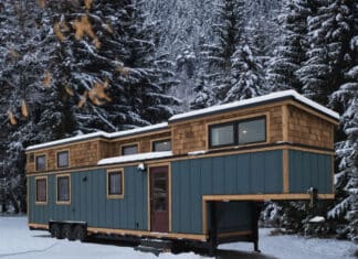 A blue tiny house with wooden trim sits on wheels in a snowy clearing, surrounded by tall, snow-covered evergreen trees and mountains in the background—a perfect winter escape for any tiny house enthusiast.