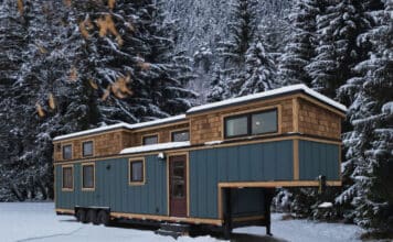 A blue tiny house with wooden trim sits on wheels in a snowy clearing, surrounded by tall, snow-covered evergreen trees and mountains in the background—a perfect winter escape for any tiny house enthusiast.