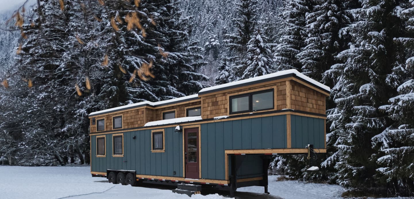 A blue tiny house with wooden trim sits on wheels in a snowy clearing, surrounded by tall, snow-covered evergreen trees and mountains in the background—a perfect winter escape for any tiny house enthusiast.