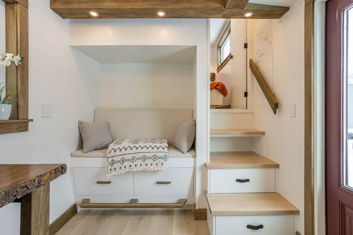 Cozy nook in a tiny house with a cushioned bench, pillows, and a patterned throw blanket. Built-in drawers beneath the bench; wooden stairs with storage lead to an upper area. Bright, modern decor with wood accents.