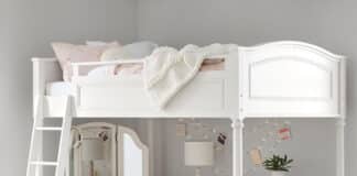 A white bed with desk below features a cozy pink chair, a corner mirror, lamp, string lights, and shelves with books and baskets. A light blanket is draped on the bed, and a blue rug brightens the floor.
