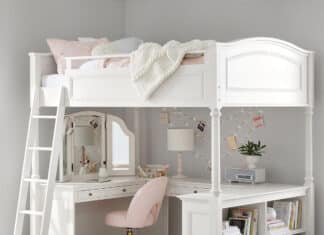 A white bed with desk below features a cozy pink chair, a corner mirror, lamp, string lights, and shelves with books and baskets. A light blanket is draped on the bed, and a blue rug brightens the floor.