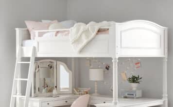 A white bed with desk below features a cozy pink chair, a corner mirror, lamp, string lights, and shelves with books and baskets. A light blanket is draped on the bed, and a blue rug brightens the floor.