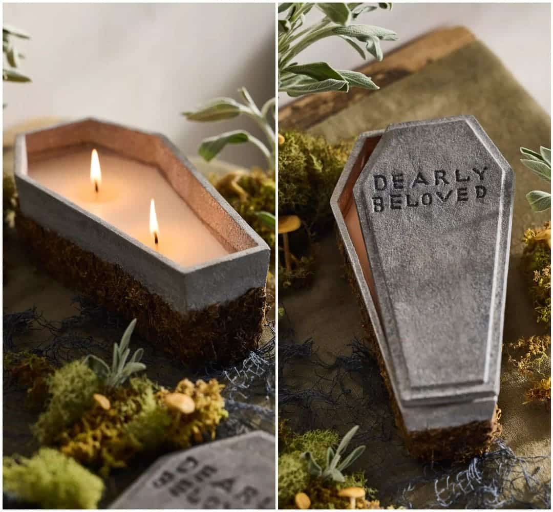 A candle shaped like a gray coffin with DEARLY BELOVED engraved on the lid, surrounded by moss and small plants. Perfect as spooky Halloween decor, this candle has two wicks and sits on a rustic surface.