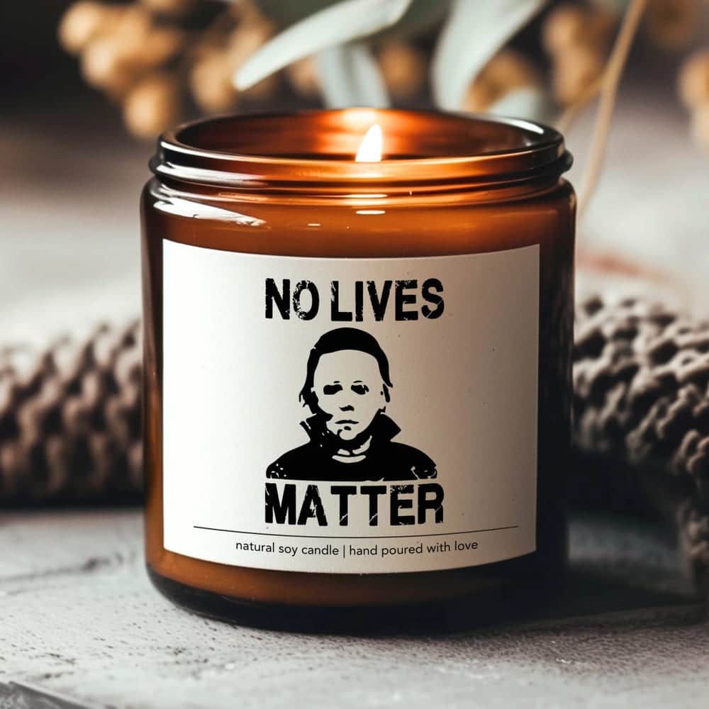 A lit brown glass candle, perfect for Halloween decor, features a label with a masked character and the words NO LIVES MATTER. The label also reads natural soy candle | hand poured with love.
