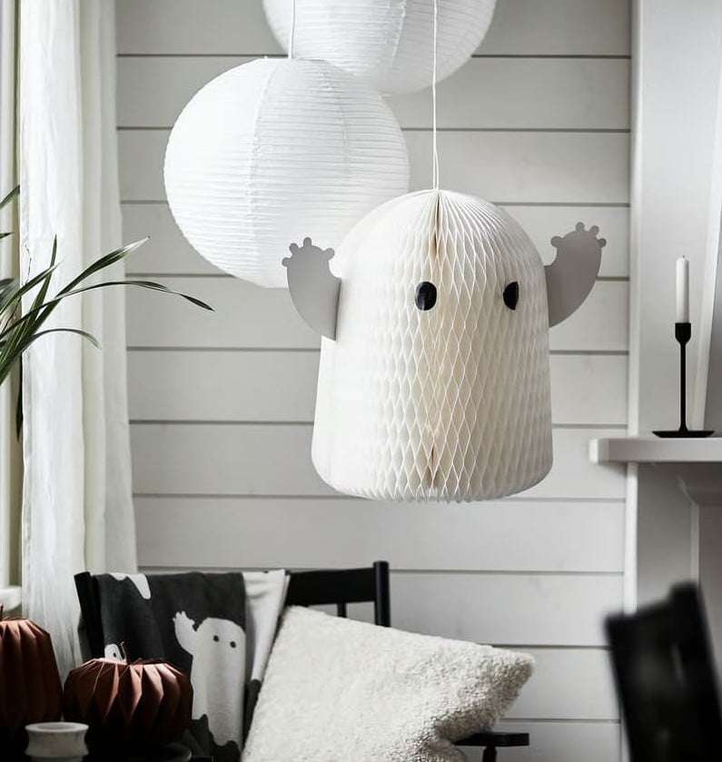 A round, white paper lantern shaped like a ghost with black eyes and raised arms hangs from the ceiling, adding a fun halloween decor touch. Two plain paper lanterns and cozy home decor are visible in the background.