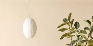 A modern wooden daybed with red and white striped cushions and two cylindrical pillows sits beside a large leafy plant, under a white pendant lamp, all set against a beige background—a cozy spot for daybed lounging.