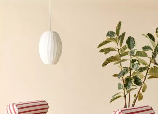 A modern wooden daybed with red and white striped cushions and two cylindrical pillows sits beside a large leafy plant, under a white pendant lamp, all set against a beige background—a cozy spot for daybed lounging.
