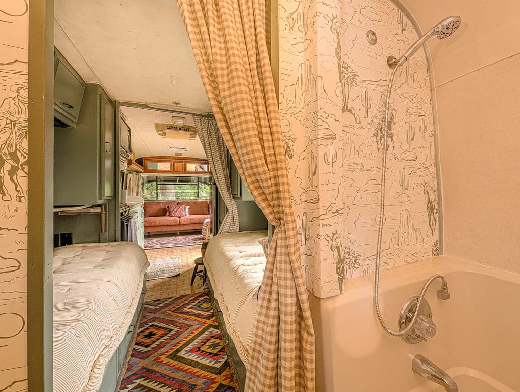 Interior of a cozy Avion camper featuring a small bathtub and shower, twin beds on either side, patterned curtains and walls, and a view into the living area with sofa, large windows, and a Southwestern-style rug on the floor.