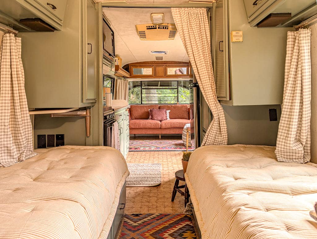The interior of this cozy Avion camper features two twin beds with beige bedding, patterned rugs on the floor, and curtains separating the sleeping area from a lounge with a red sofa and large windows in the background.
