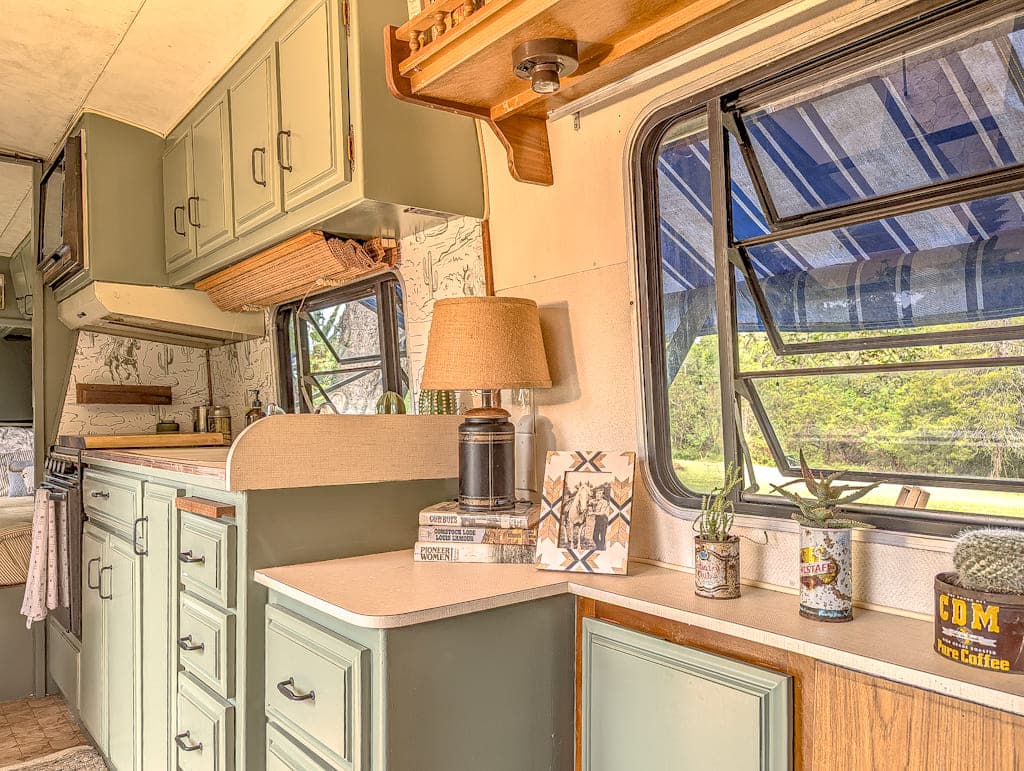 Interior of a cozy Avion camper kitchen with green cabinets, open window, countertop decor including a lamp, books, small plants, and a tin can labeled “CDM;” patterned wallpaper and an outdoor awning visible through the window.