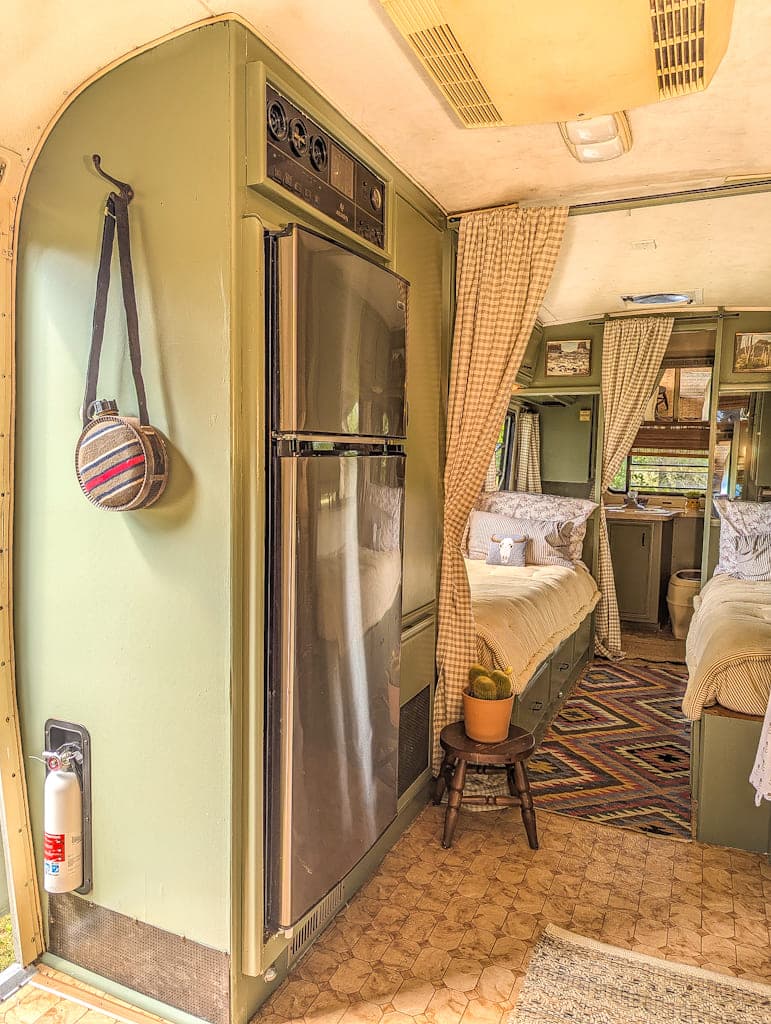 A cozy, vintage Avion camper interior with a fridge, hanging bag, potted plant on a stool, curtained sleeping area with two beds, patterned rug, and a fire extinguisher by the door. Warm, natural light fills the space.