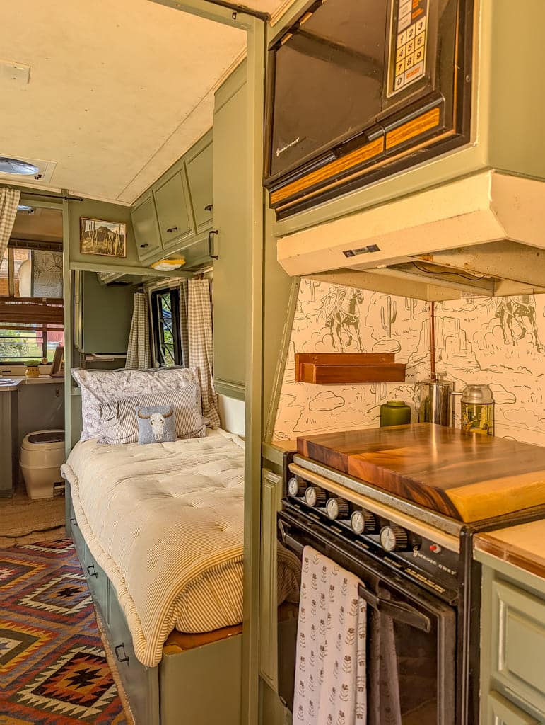 A cozy Avion camper interior featuring a neatly made bed, sage green cabinets, a patterned rug, and a compact kitchen with a stove, oven, cutting board, and microwave—all bathed in natural light with desert-themed decor.
