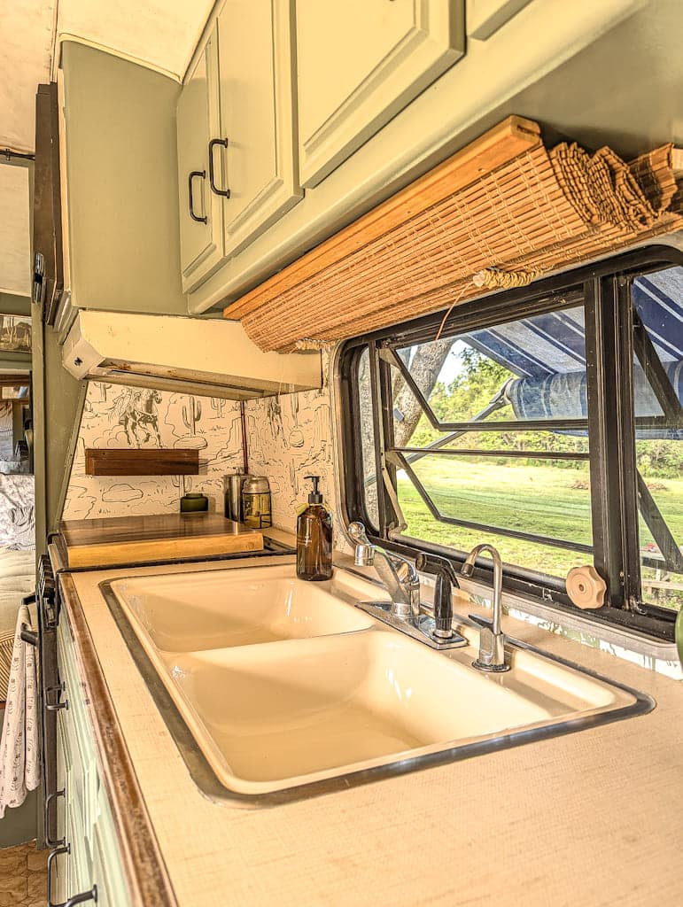 A bright Avion camper kitchen features a double sink, faucet, soap dispenser, small appliances, green cabinets, a window with a bamboo shade, and scenic greenery visible outside.