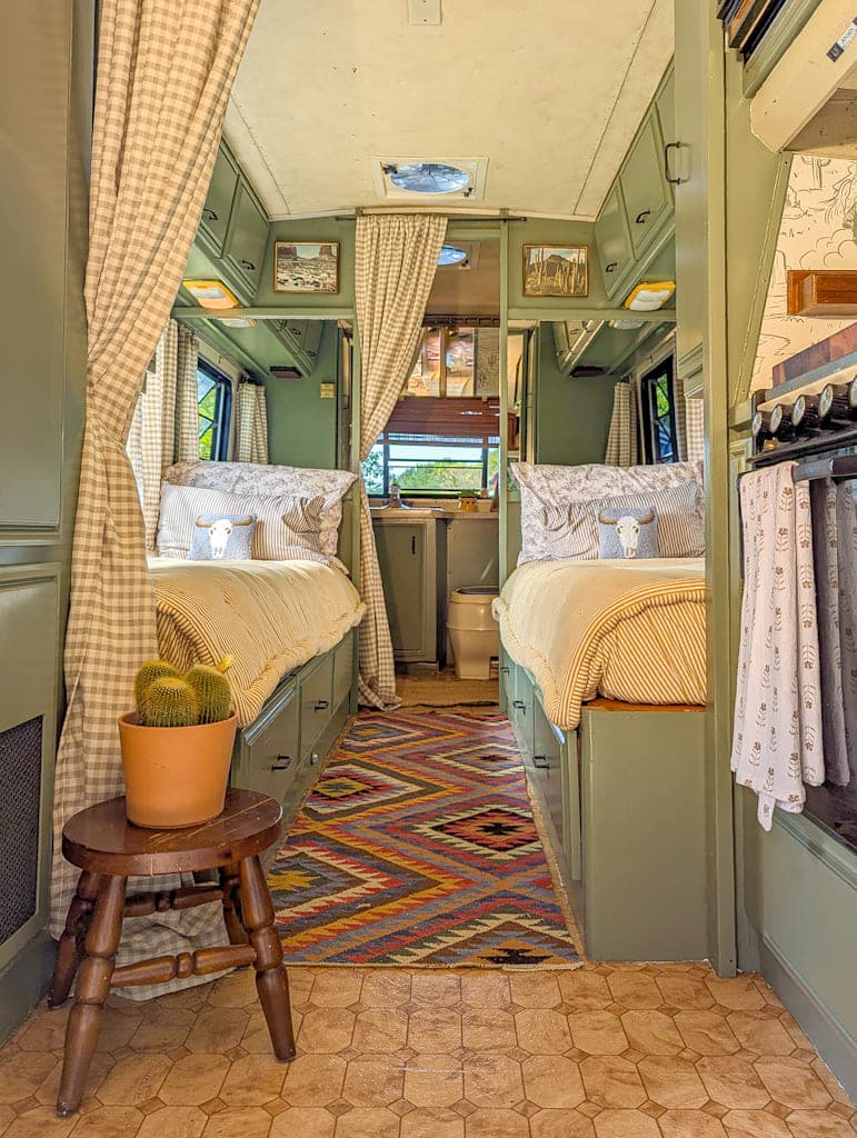 Interior of a cozy, renovated Avion camper bus with two twin beds, neutral bedding, patterned rug, potted cactus on a stool, curtains for privacy, and a kitchenette on the right. Natural light streams in from the windows.