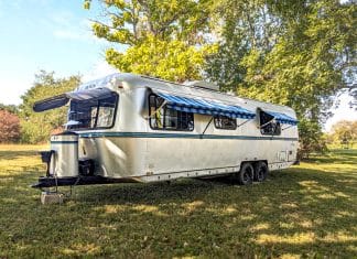 A silver vintage Avion camper with blue-striped awnings is parked on grass under shady trees on a sunny day. The trailer has two axles and several windows open for ventilation.
