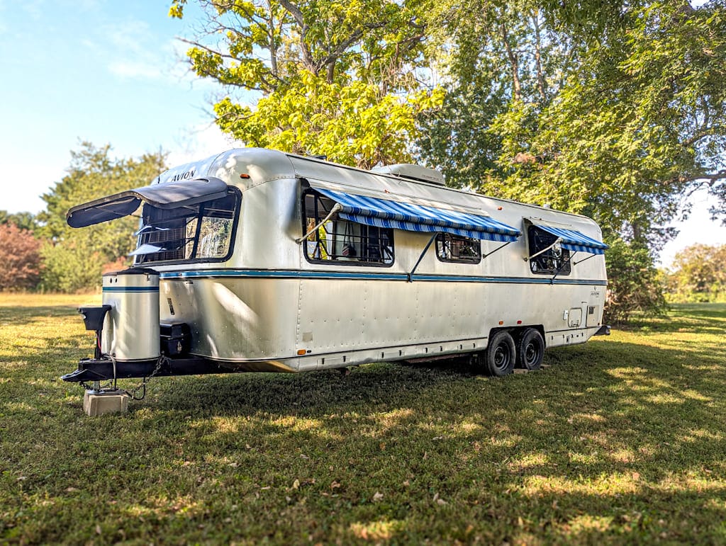 A silver vintage Avion camper with blue-striped awnings is parked on grass under shady trees on a sunny day. The trailer has two axles and several windows open for ventilation.