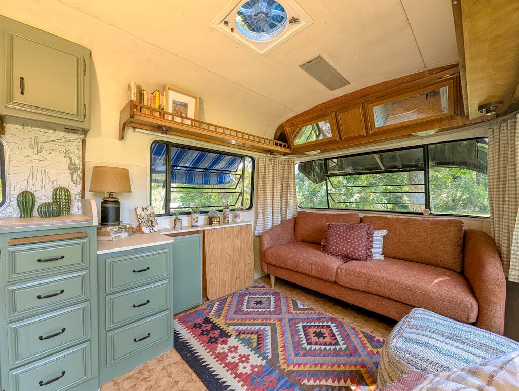 Cozy Avion camper interior with green cabinets, a countertop, lamp, and decor on the left. A brown sofa with cushions sits on a colorful patterned rug, with large windows letting in natural light and trees visible outside.