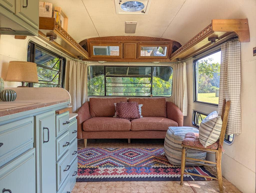A cozy renovated Avion camper interior featuring a rust-colored sofa, patterned rug, ottoman, wooden chair with cushions, and large windows with checkered curtains letting in sunlight. Painted cabinets are on the left.