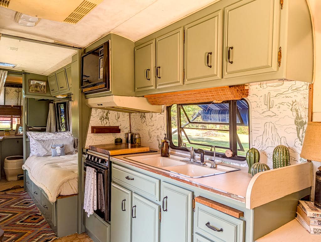 A cozy Avion camper interior featuring sage green cabinets, a kitchen with a double sink, stove, and microwave, desert-themed wallpaper, a bed in the back, and sunlight streaming through the window.