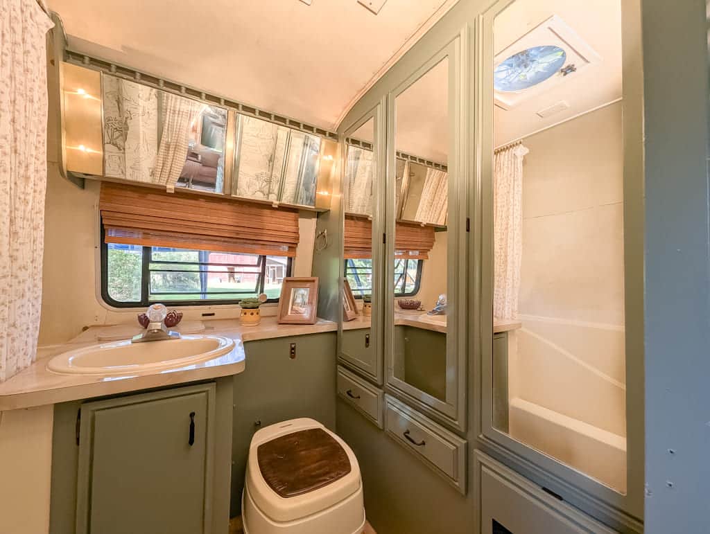A cozy Avion camper bathroom with a sink, mirror cabinets, toilet, wooden blinds on windows, and a small bathtub with shower curtain. Light green cabinetry and soft lighting create a warm, inviting atmosphere.