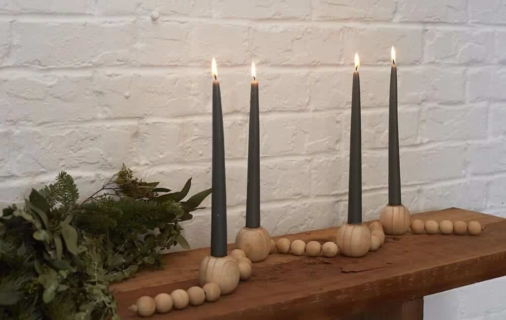 Four lit black taper candles in round wooden candle holders stand upright on a wooden surface, accompanied by a green leafy arrangement and a string of wooden beads, all set against a white brick wall.