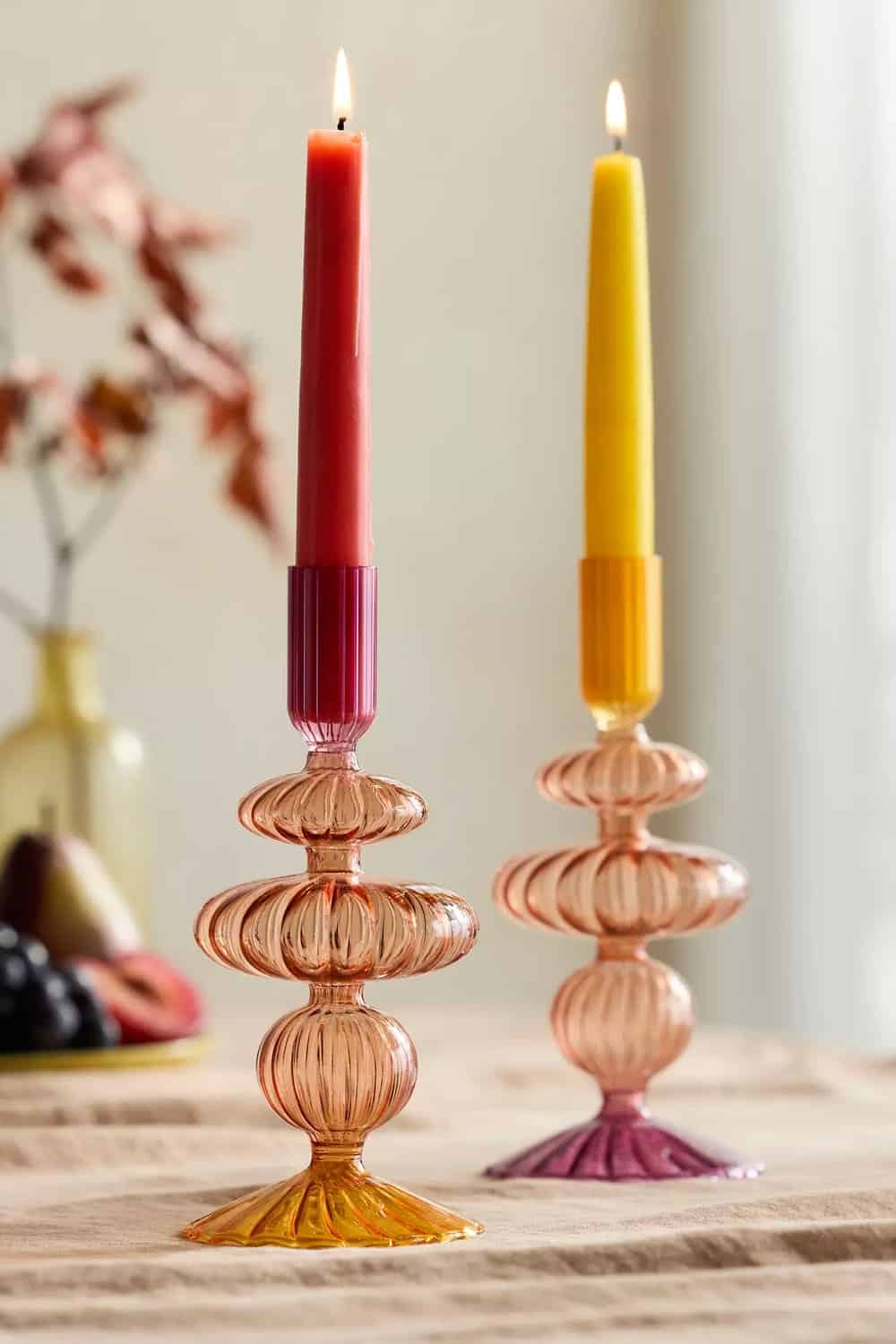 Two colorful glass candle holders with textured bases hold lit candles—one red and one yellow—on a table. Blurred fruit and a vase with leaves add visual interest in the background.