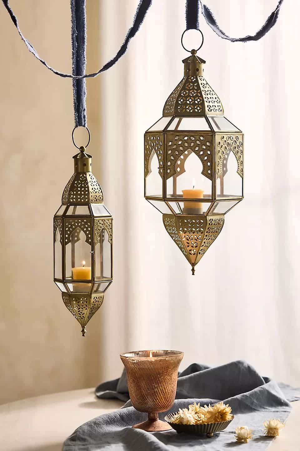 Two ornate brass lanterns with lit candles hang from blue ribbons. Below, a glowing candle sits in a textured candle holder next to a small bowl of white flowers on a draped blue cloth.