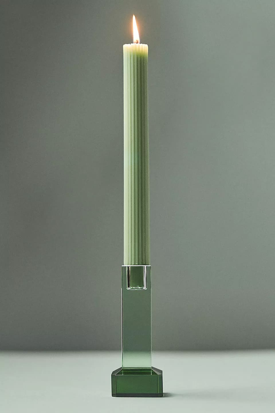 A lit cream-colored ribbed candle stands upright in a striking green glass candle holder against a plain gray background.