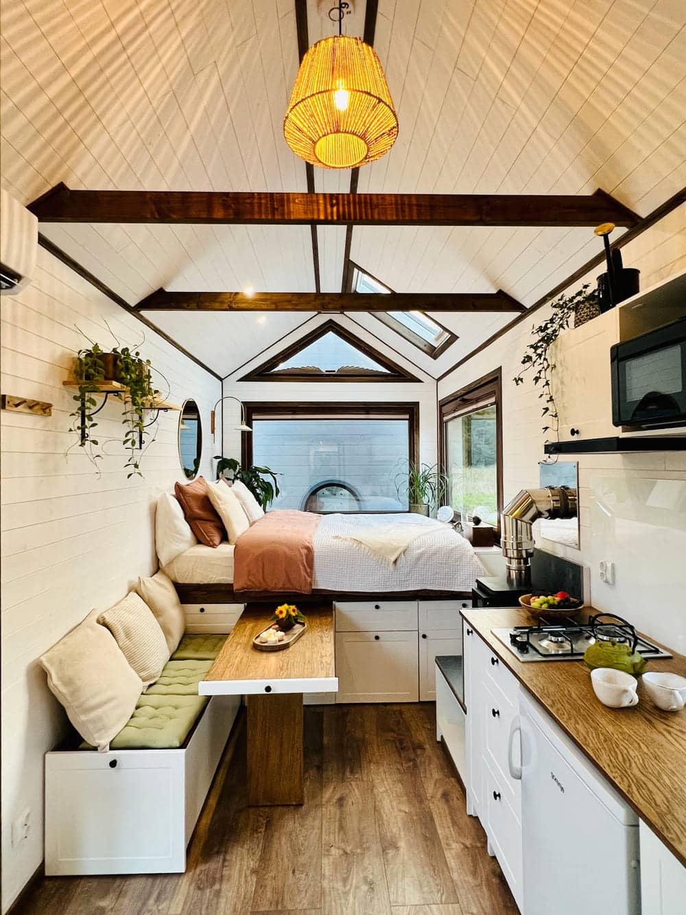 A cozy, well-lit tiny house interior features a bed with blush bedding, a cushioned bench dining area, a small kitchen with wood countertops, lush plants, and large windows that fill the tiny house with natural light.