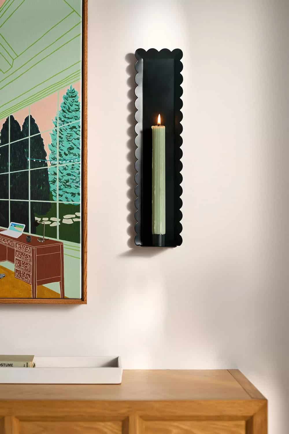 A scalloped black candle holder wall sconce with a tall green candle is mounted on a white wall next to a colorful framed artwork above a light wood cabinet.