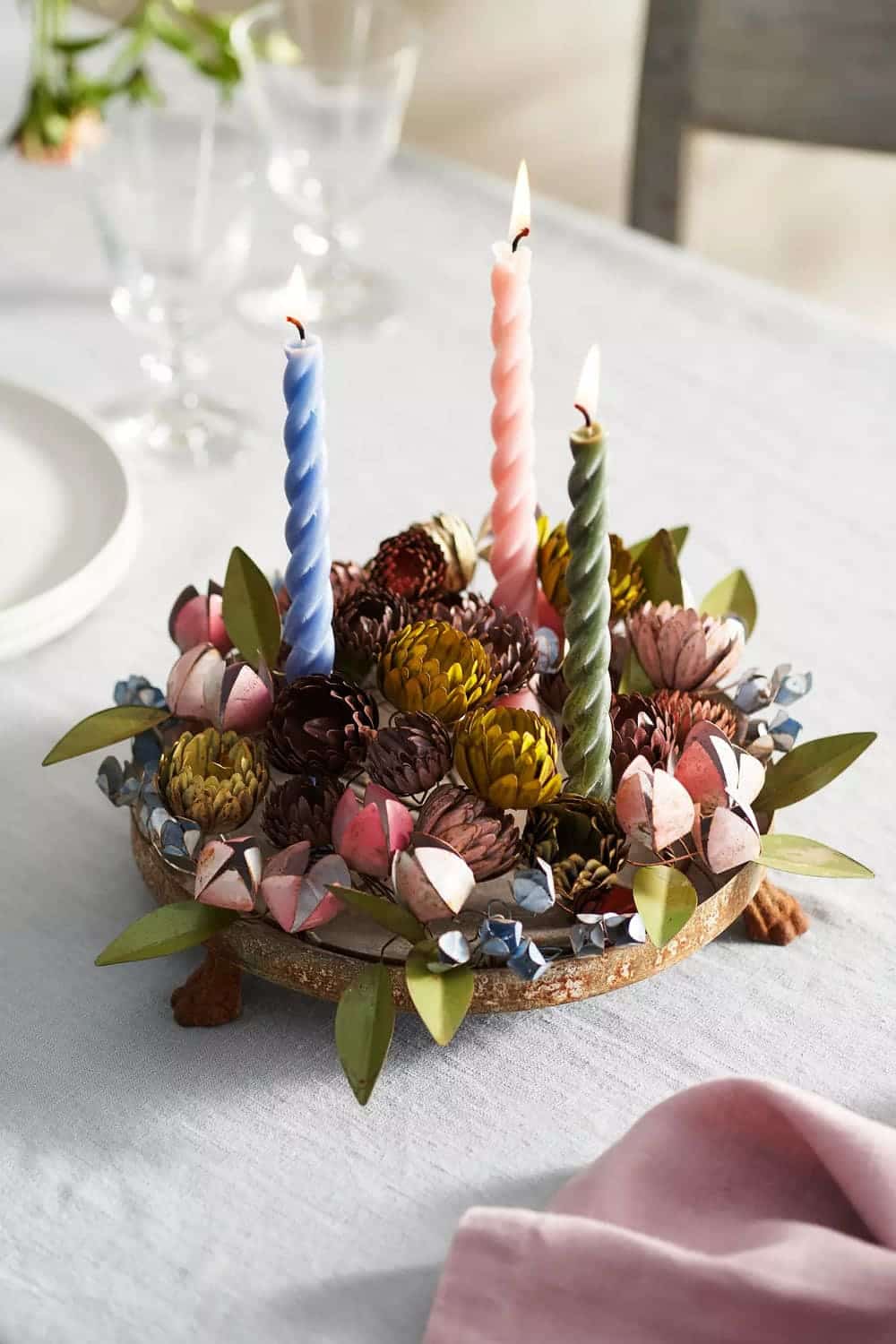 A floral centerpiece with pink, purple, and green flowers surrounds three lit spiral candles—blue, pink, and green—in a stylish candle holder on a wooden base. The display sits on a light gray tablecloth near plates and glasses.