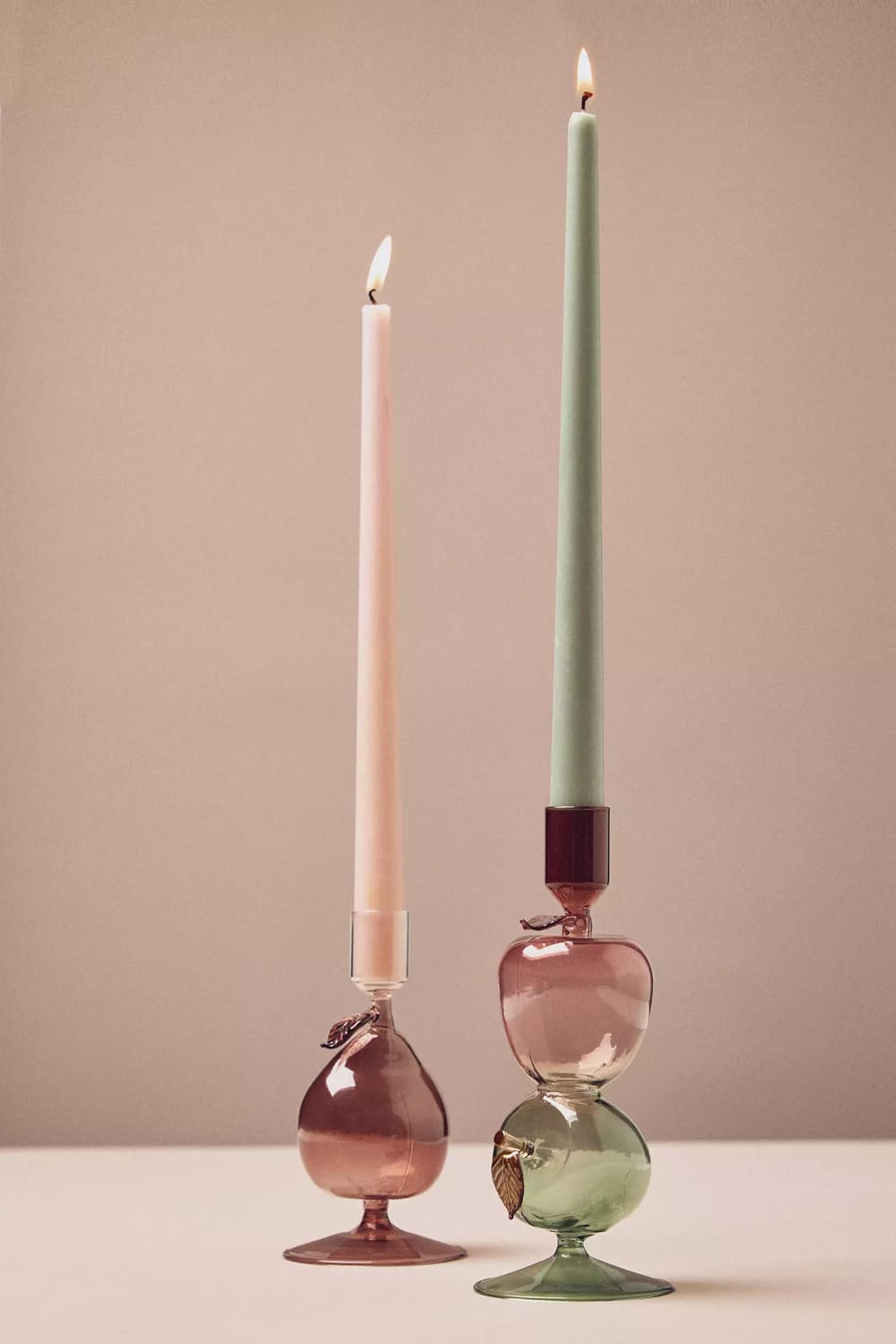 Two glass candle holders, each with an artistic, rounded base in pink and green tones, hold tall, lit taper candles—one pale pink and one mint green—against a neutral background.