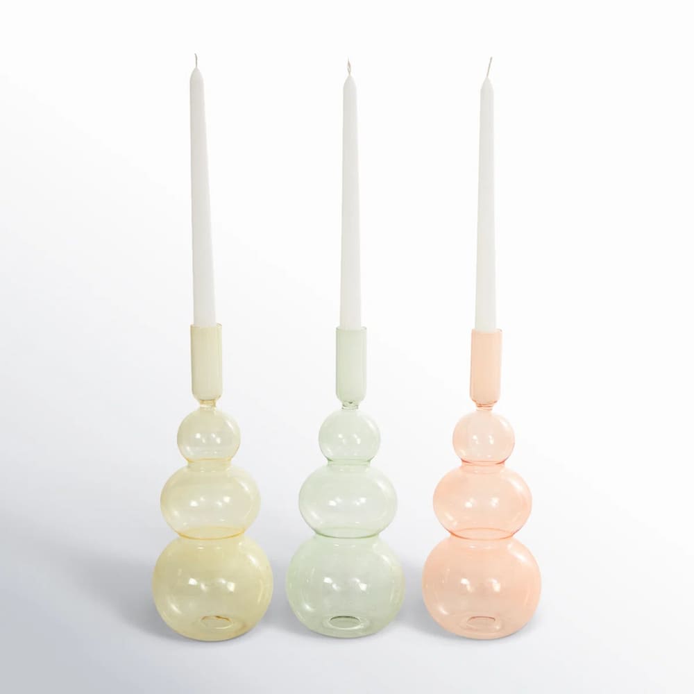 Three pastel-colored candle holders, each with stacked spherical shapes, hold tall white taper candles. The glass candle holders—yellow, green, and pink—are arranged side by side on a plain background.