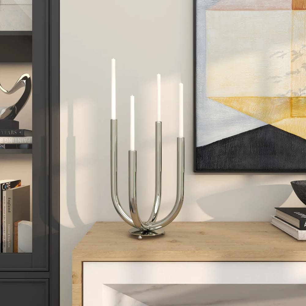 A modern silver candle holder with four lit white candles sits on a light wooden surface beside a black bookshelf and a framed abstract painting on the wall.