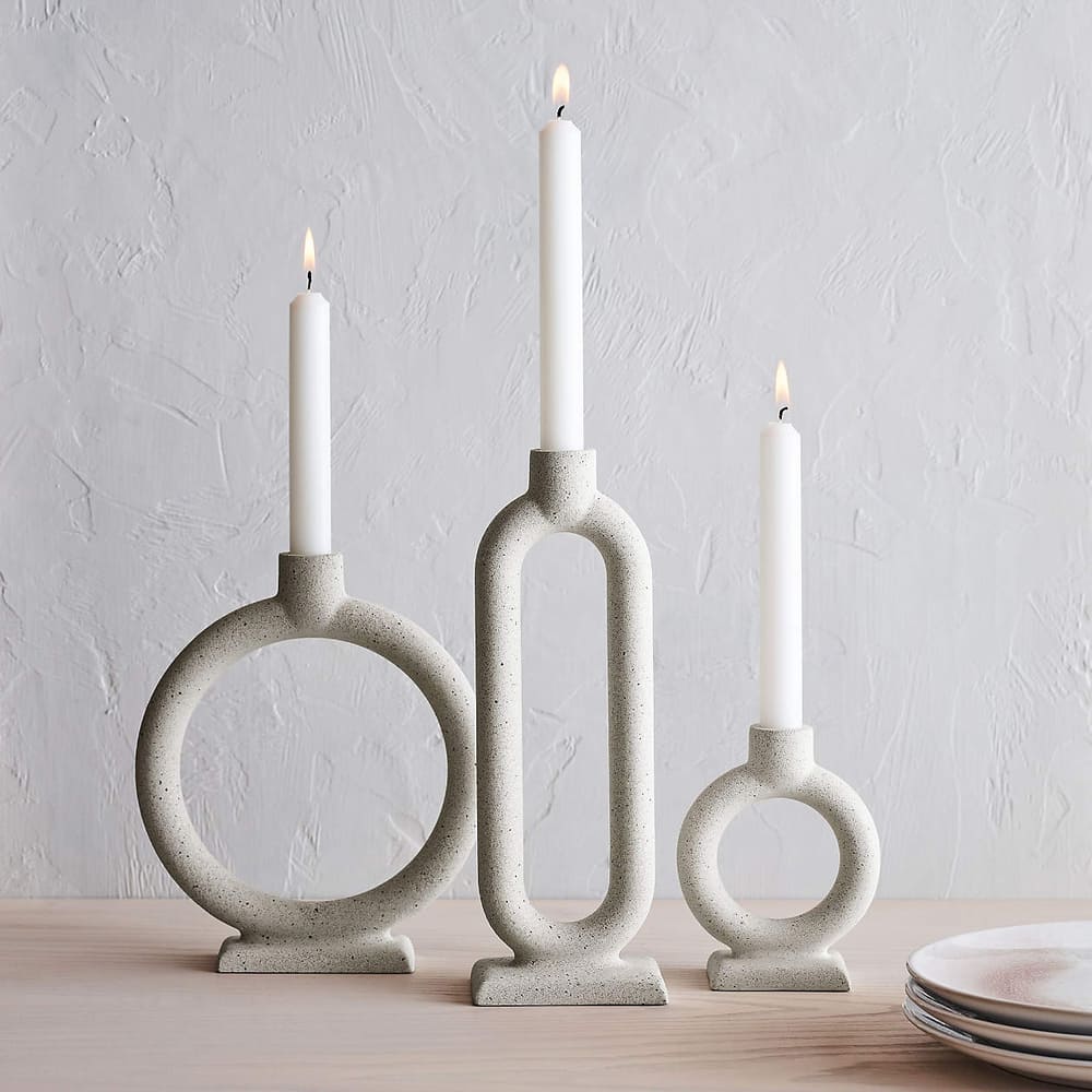 Three modern, sculptural candle holders in light gray stone hold white taper candles of varying heights on a wooden surface against a textured light gray wall. Candle holder silhouettes stand out, with plates partially visible on the right.