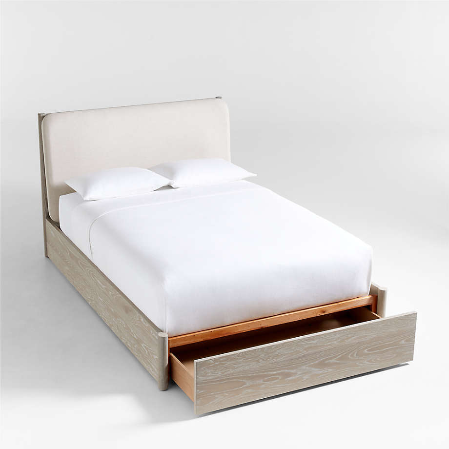 A modern storage bed featuring light wood, a fabric-upholstered headboard, crisp white bedding, two pillows, and a spacious storage drawer pulled out at the foot of the bed.