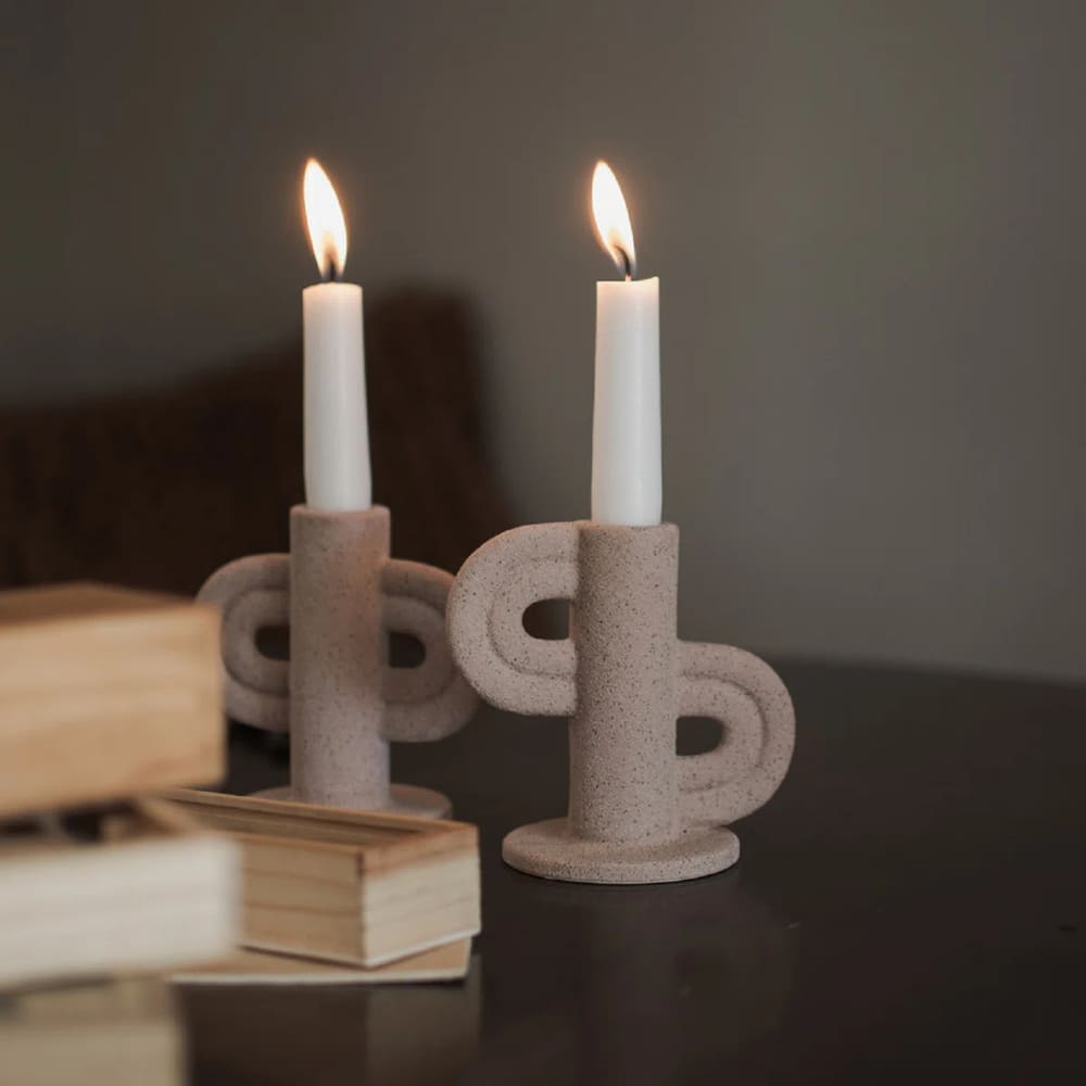 Two lit white candles in sculptural beige candle holders sit on a dark surface next to a stack of wooden items. The scene exudes a warm, minimalistic, and cozy atmosphere.