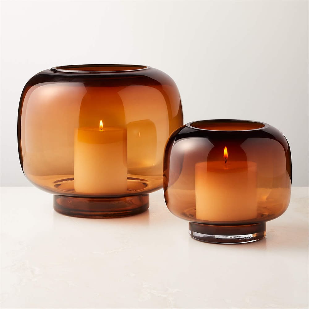 Two lit pillar candles rest inside round, translucent candle holders of amber glass in varying sizes, displayed on a white surface against a neutral background.